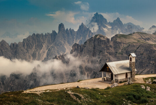 View of a serene chapel clinging to the mountainside, shrouded in ethereal clouds and majestic, jagged peaks, Misurina, Veneto, Italy.