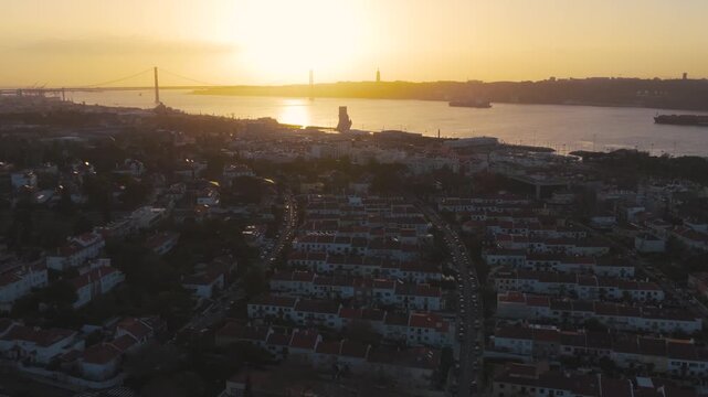 Drone view over Lisbon at sunset with the 25 de Abril Bridge spanning the Tagus River and Christ the King monument visible in the distance. Warm golden light over the Portuguese capital skyline.
