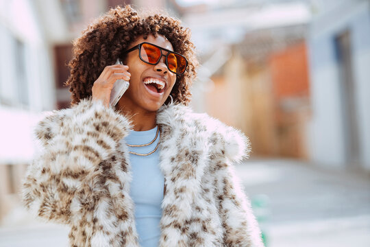 Happy young afro woman talking on smartphone, wearing sunglasses and a leopard print coat walking outdoors at urban street.
