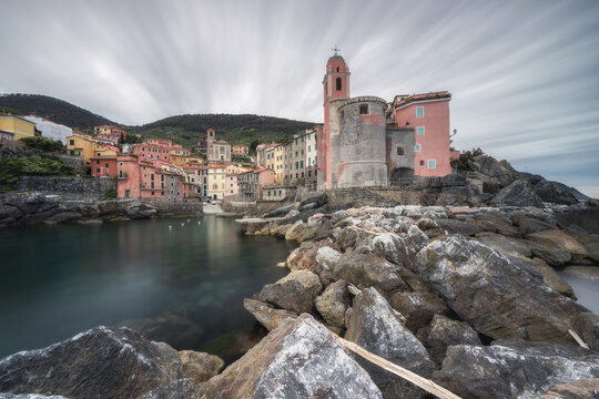 View of the colorful buildings and the church standing on the rocks overlooking the sea, the sky streaked with clouds, Tellaro, Liguria, Italy.