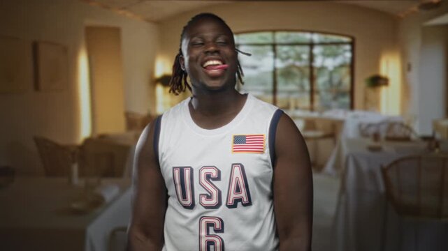 Man smiling and laughing with bare shoulders visible, wearing usa basketball jersey with flag patch in a restaurant building, facing camera; joy celebration.