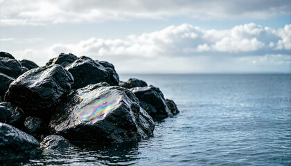 Fototapeta premium Oil spill rainbow sheen on black rocks by the ocean. Marine pollution and environmental disaster concept