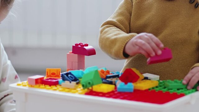 Two children build something using colorful blocks on a table. They focus on stacking pieces while surrounded by more blocks scattered around them