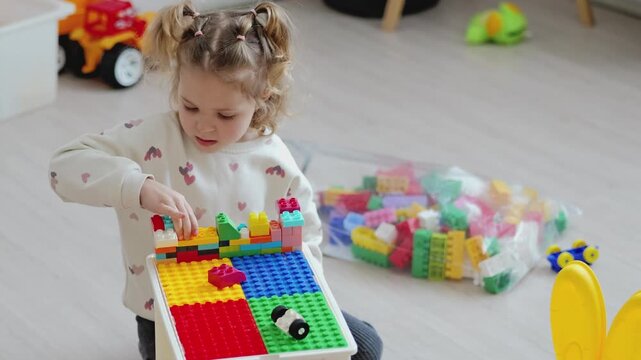 A young child constructs a design using colorful building blocks in a play area filled with toys. The space is well lit with natural light