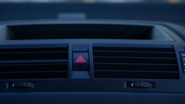 Close-up view reveals the dashboard features of a modern car, highlighting air vents, control switches, and an emergency signal button. The alarm button is on and flashing.