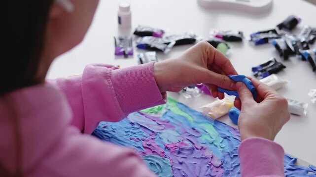 Individual mixes colors and applies paint on a canvas in a bright room. Art supplies and paint tubes are scattered on the table nearby