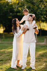 Fototapeta premium Happy Young Family Outdoors: Father Carrying Son on Shoulders While Mother Smiles in Park