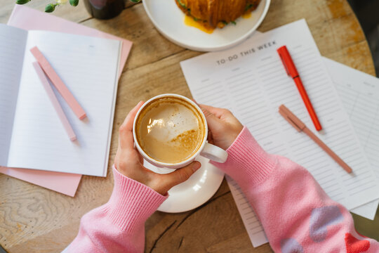 Overhead view of a woman sitting at a table with a blank to do list drinking a cappuccino and eating a savoury croissant