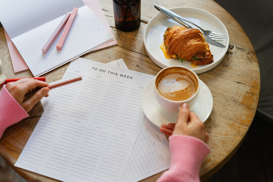 Overhead view of a woman sitting at a table with a blank to do list drinking a cappuccino and eating a savoury croissant