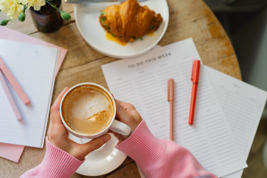 Overhead view of a woman sitting at a table with a blank to do list drinking a cappuccino and eating a savoury croissant