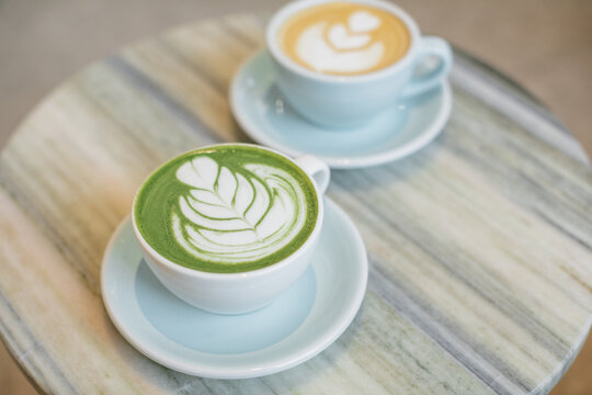 Close-up of a hot matcha latte and a cappuccino with floral latte art decoration on a table