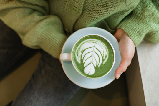 Close-up overhead view of a woman in a green cardigan sitting in a cafe drinking a hot matcha latte