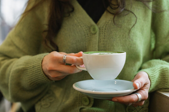 Close-up front view of a woman in a green cardigan sitting in a cafe drinking a hot matcha latte