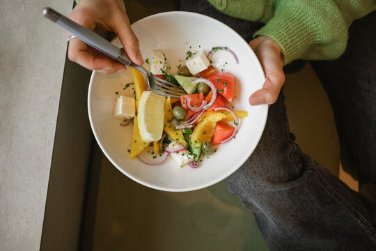 Overhead view of a woman sitting at a table eating a fresh Greek salad with feta cheese, tomato, olives, onions and bell peppers