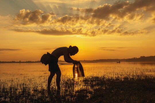 Side view silhouette of a fisherman standing in a river holding a fishing trap at sunrise, Thailand