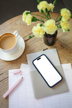Overhead view of a mobile phone with a blank screen on a weekly planner, a stack of papers with a cappuccino, highlighter pen and bunch of flowers in a vase on a wooden table