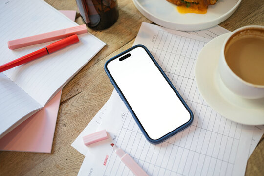 Overhead view of a mobile phone with a blank screen on a weekly planner with a highlighter pen, cup of coffee, savoury cheese and salad croissant on a wooden table