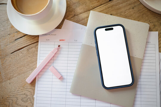 Overhead view of a mobile phone with a blank screen on a weekly planner, highlighter pen, notebook and a cappuccino on a wooden table
