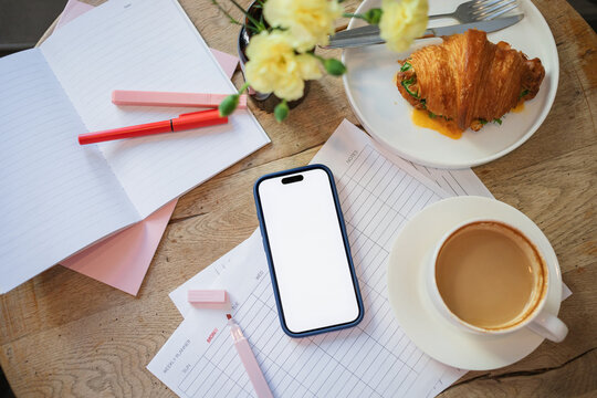 Overhead view of a mobile phone with a blank screen on a weekly planner with a highlighter pen, cup of coffee, savoury cheese and salad croissant and bunch of flowers on a wooden table