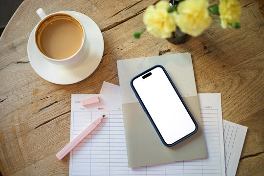 Overhead view of a mobile phone with a blank screen on a weekly planner with a highlighter pen, cup of coffee and bunch of flowers on a wooden table