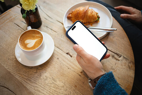Close-up of a man sitting at a table with a cappuccino and savoury croissant holding a mobile phone with a blank screen