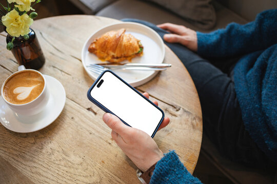 Close-up of a man sitting at a table with a cappuccino and savoury croissant holding a mobile phone with a blank screen