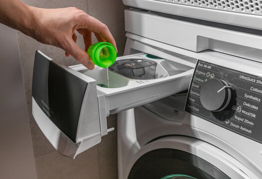 Woman pouring liquid detergent into washing machine dispenser tray. Adult female holding green cap above appliance drawer in laundry room. Domestic washing concept for home, household,
