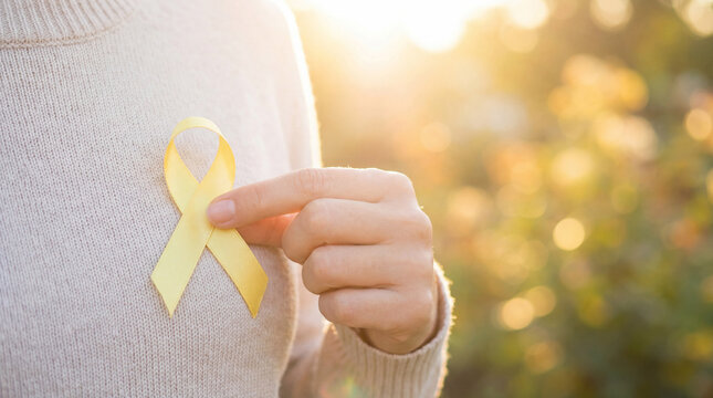 Close-up of a  hand holding a yellow awareness  ribbon. Soft sunlight and a blurred backround.