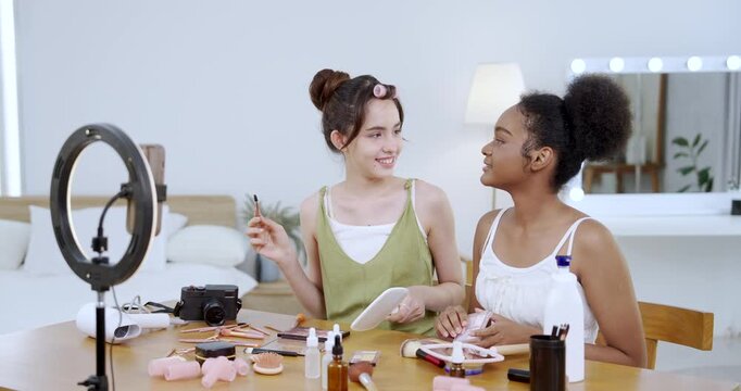 asian teenage girl holding makeup palette while smiling with african american teenage girl during beauty tutorial content shoot at home showing influencer lifestyle setup with cosmetic tools on table