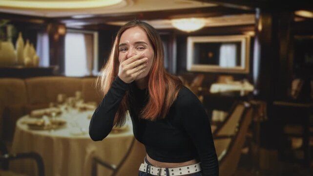 Young redhead woman with hand covering mouth, laughing and leaning forward inside a dining building; surprise playful joy.