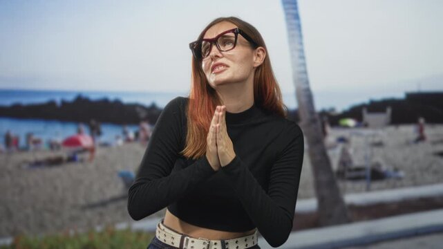 Woman with hands pressed together in a praying gesture on a street near the beach, wearing glasses and black turtleneck, freckled young; hope reflection.