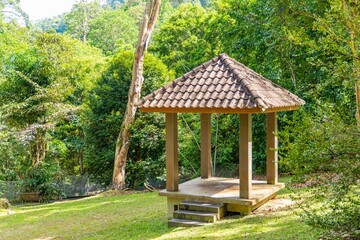 Small concrete pavilion with tiled roof situated on green grassy hill, surrounded by dense tropical forest trees and vegetation in public park.