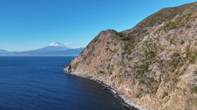 Breathtaking aerial footage of the rugged coastline and cliffs of Ita in Numazu, Izu Peninsula, featuring the majestic Mt. Fuji visible across the deep blue Suruga Bay on a sunny winter day.
