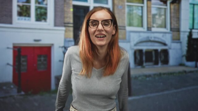 Young redhead woman with glasses, mouth open shouting and leaning forward with one hand cupped near face on street; playful spontaneity.