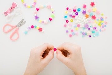 Naklejka premium Girl hands making colorful bracelet on white table background. Creating different beaded jewellery. Little child handcraft. Closeup. Point of view shot. Top down view.