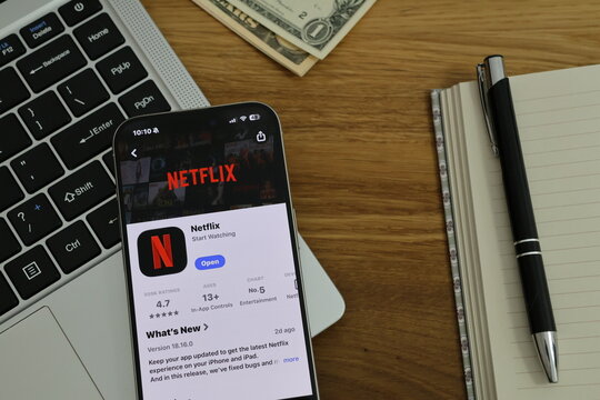 Kiel, Germany - 02-25-2026: A smartphone displaying the Netflix video streaming app for movies and series in App Store displayed on smartphone placed on top of laptop on wooden des