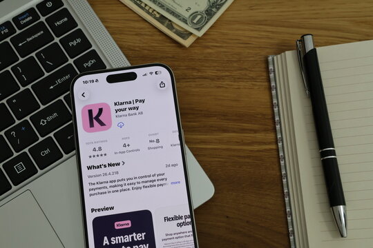 Kiel, Germany - 02-25-2026: A smartphone displaying the Klarna buy now pay later and shopping payments app in App Store displayed on smartphone placed on top of laptop on wooden desk