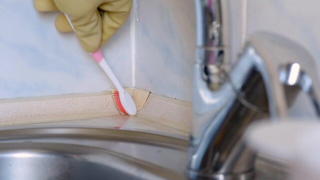 Gloved hand scrubbing sink grout near faucet, closeup of toothbrush bristles working into tile joint to remove brown residue and mold, yellow rubber glove, stainless faucet
