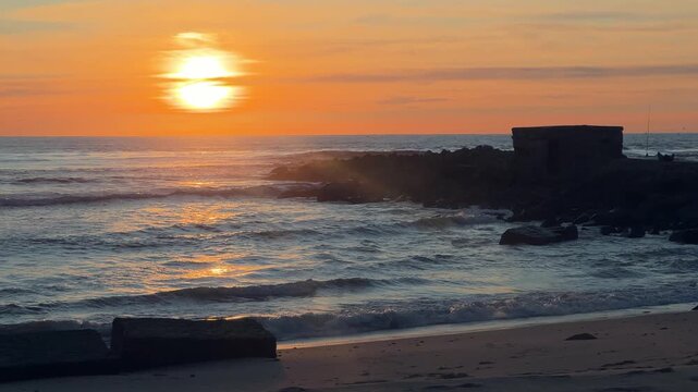 Static shot of sunset over ocean waves breaking against stone breakwater on sandy beach in Barbate, Cadiz, Spain.
