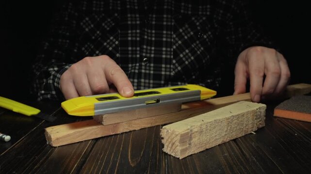 Individual using a yellow level tool to measure wooden planks on a dark wooden table, showcasing precise alignment and craftsmanship in a workshop setting