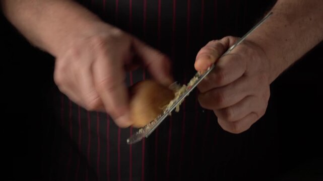 Hands of a male chef grating a piece of vegetable using a stainless steel grater, showcasing the continuous motion of the grating process in a dark kitchen setting