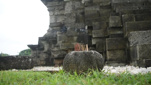 Incense sticks burning in a stone container near a temple, with soft smoke billowing into the air. 