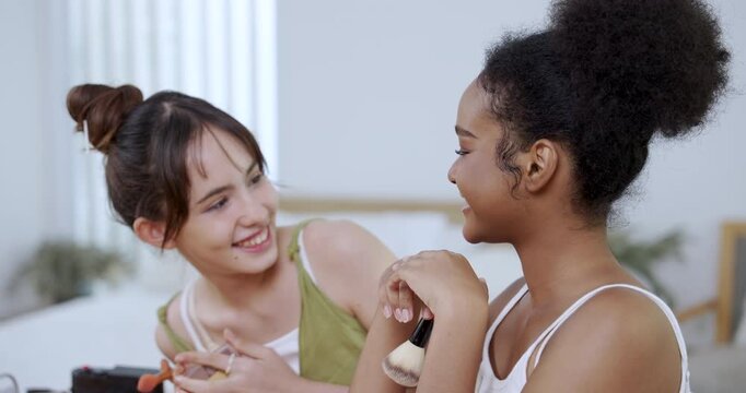 african american teenage girl holding makeup brush while smiling with asian teenage friend during slowmotion beauty tutorial session at home studio showing joy in cosmetic influencer lifestyle content