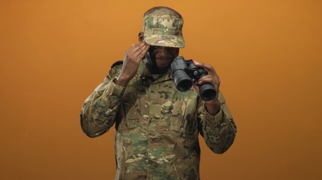 Man soldier holding binoculars and touching cap while scanning ahead in studio, camouflage uniform visible; duty vigilance.