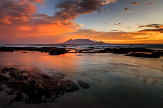 View of fiery sunset paints the sky above the tranquil sea, reflecting on the wet rocks, with Table Mountain silhouetted in the distance, Cape Town, Western Cape, South Africa.