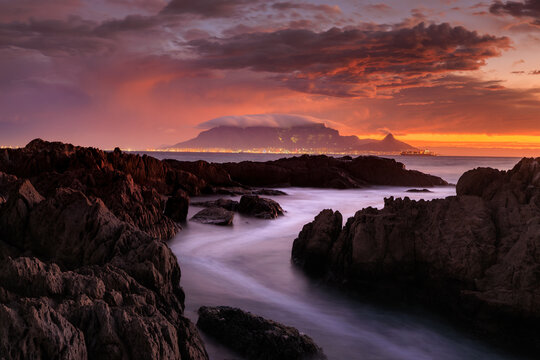 View of rugged, dark rocks frame the serene, smooth water leading to the distant, glowing Table Mountain under a dramatic, fiery sky, Cape Town, Western Cape, South Africa.