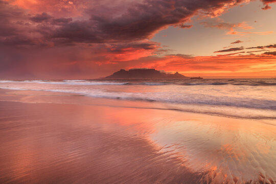 View of fiery sunset paints the sky and reflects on the wet sand of the beach, with a silhouette of a distant mountain, Cape Town, Western Cape, South Africa.