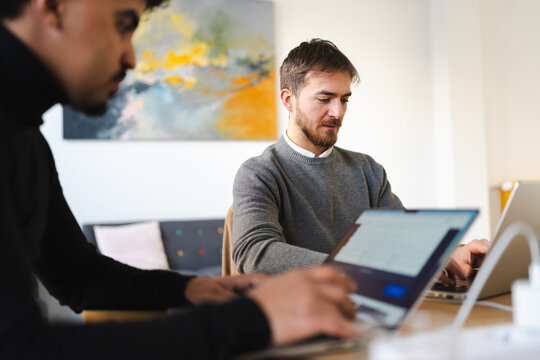 Two men collaborate on laptops in a modern office setting, focusing on their screens and tasks.