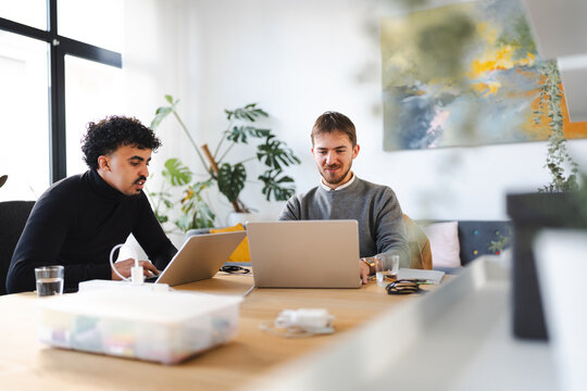 Two men collaborate on laptops at a wooden table in a bright, modern office space with plants and artwork.