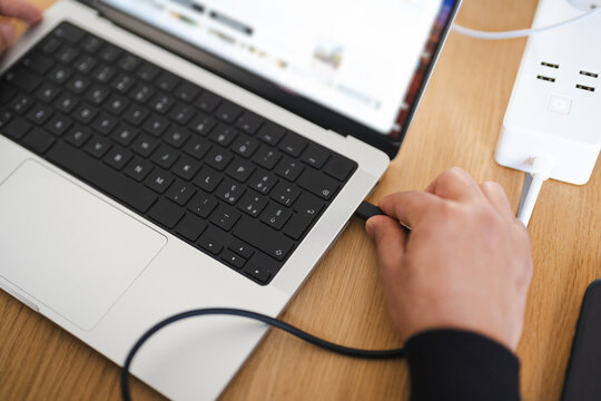 A person connects a cable to a laptop, charging it from a power strip with multiple USB ports.
